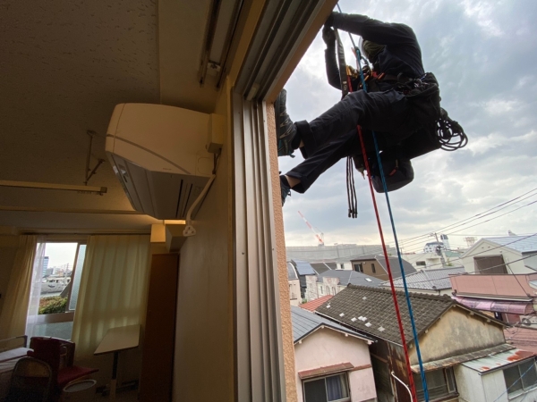 病院の室内から見た、窓の外でロープアクセスによる作業を行う職人